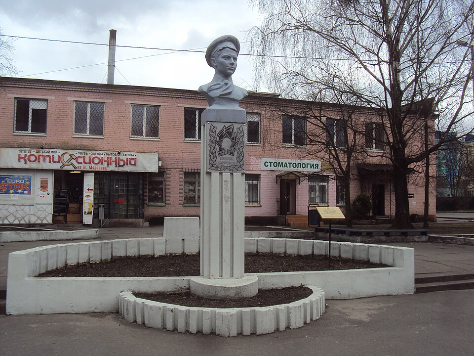 Grey stone bust of a young man in a peaked cap on a tall square pedestal at a street corner, with shops and bare trees behind, in Kaliningrad