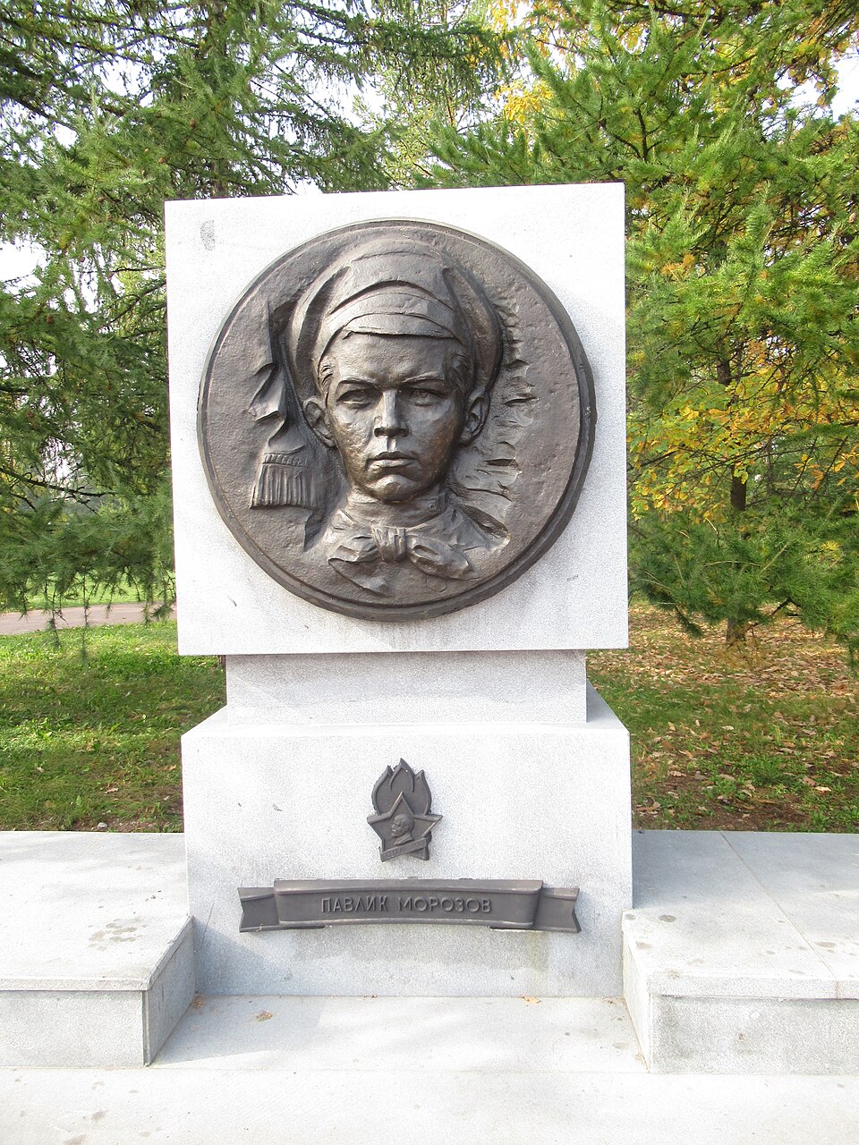 Bronze relief plaque on a stone pedestal, showing Pavlik Morozov's head and shoulders in a Pioneer scarf beside a Pioneer flame emblem, with his name cast below, in a public square in Chelyabinsk