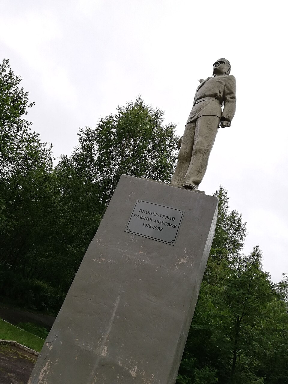 Full-length grey statue of a young man in an open jacket, looking upward with one arm raised, on a tall plinth reading in Russian "Pioneer-Hero Pavlik Morozov, 1918-1932," set among birch trees in a park