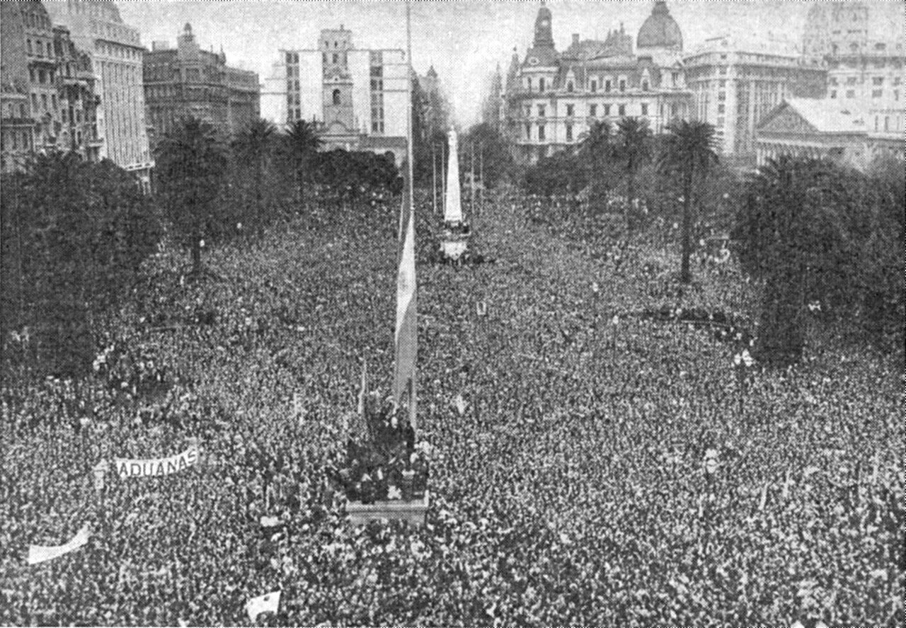 Aerial photograph of a dense crowd filling the Plaza de Mayo, Buenos Aires, around a flagpole, with the Pirámide de Mayo and buildings behind