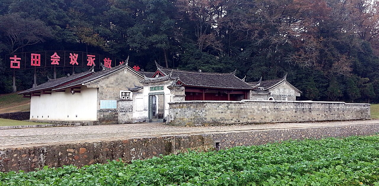 Photograph of the Gutian Congress site in Shanghang County, Fujian: a low grey-brick compound with tiled roofs below a wooded hill, and a banner of red characters naming the Gutian Conference