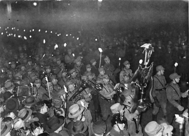Night photograph of a dense column of marchers in caps and uniform carrying torches, with a brass band and a standard, and spectators in hats in the foreground