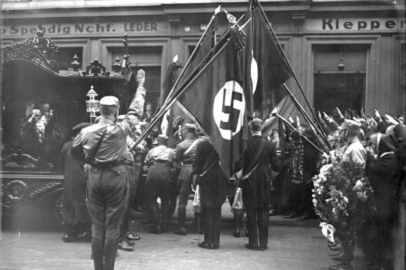 Photograph of men in uniform and civilian dress beside a hearse, raising their right arms in salute beneath dipped flags, one bearing a swastika, with a wreath at right