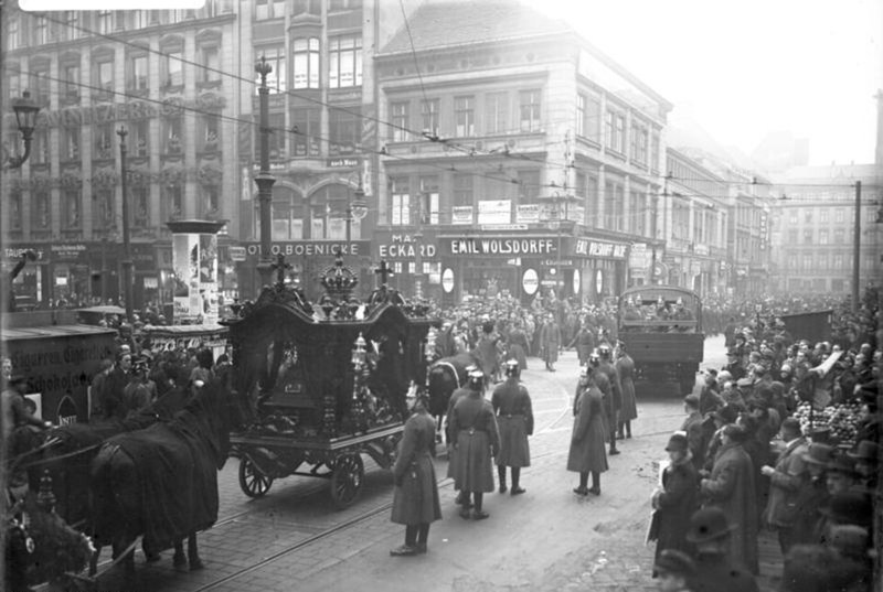 Photograph of a horse-drawn hearse in a Berlin street lined with spectators, police in the roadway and a lorry behind