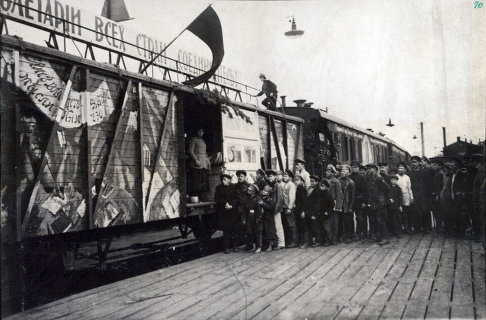 Crowd of men, women and boys standing beside a painted railway wagon, with a red flag and a banner above the roofline