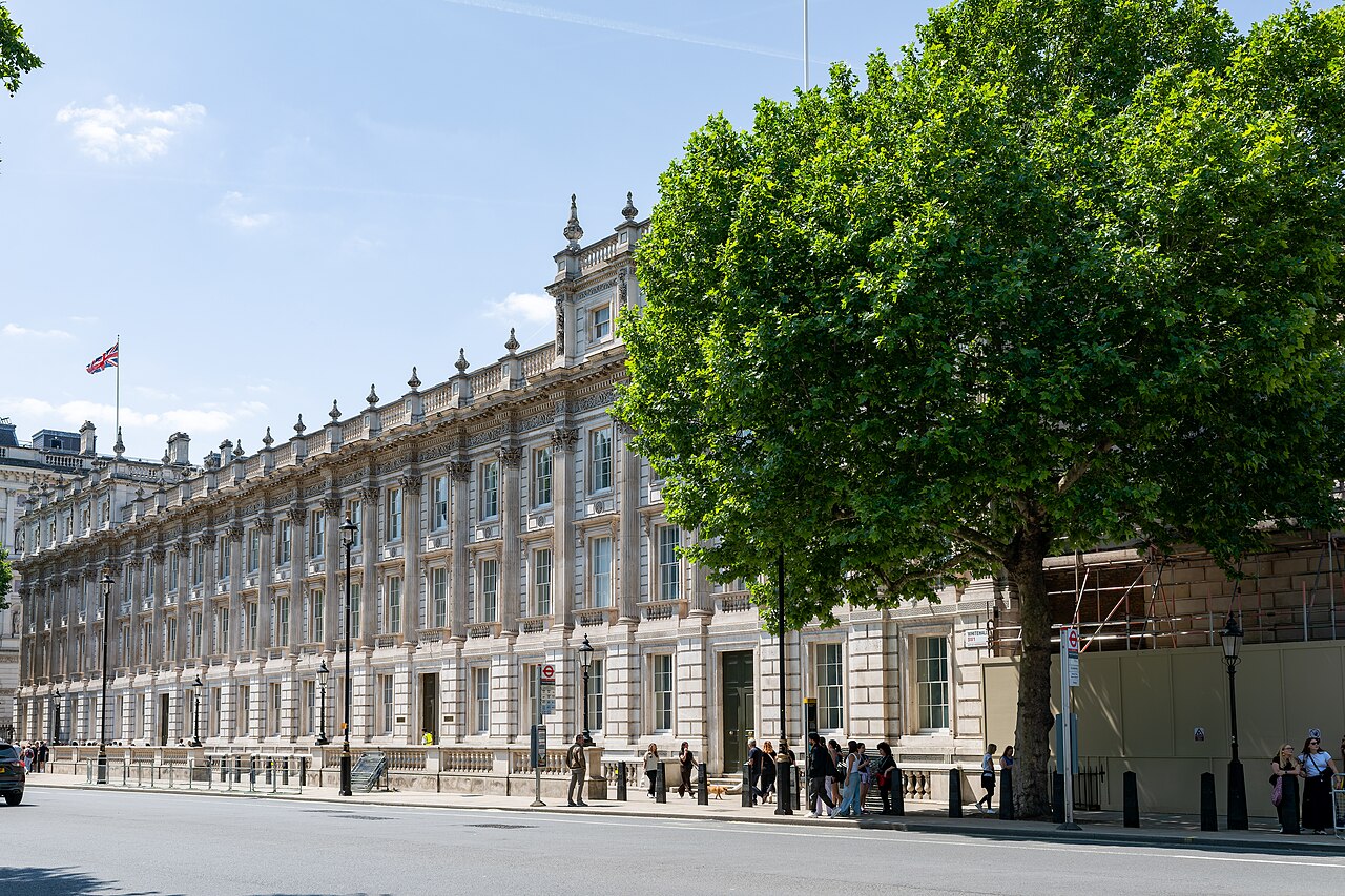 A long stone government building on Whitehall with rows of tall windows and a Union flag on the roof, beside a large plane tree and pedestrians
