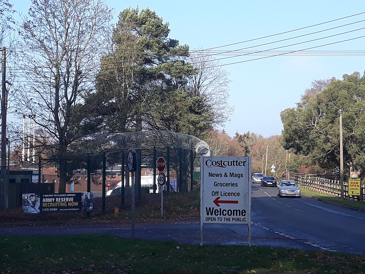 A road junction with a barracks entrance behind razor wire, a banner reading Army Reserve Recruiting Now for 73 Military Intelligence Company, and a shop sign in the foreground
