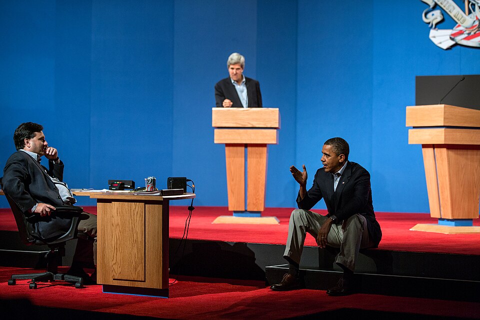 President Barack Obama on a debate stage beside a table where a seated aide rests his chin on his hand; a man stands at a lectern in the background