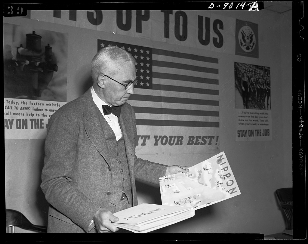 Elmer Davis, in glasses and a bow tie, looks down at bound volumes labeled "Germany" and "Nippon" in front of wall posters reading "It's up to us" and "Stay on the job"