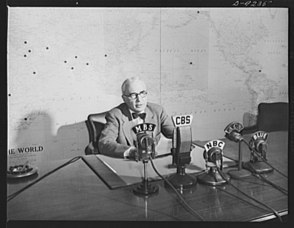 Elmer Davis seated at a desk before four radio-network microphones, with a wall map behind him