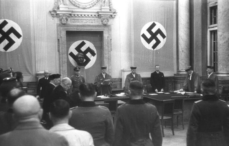 Volksgerichtshof courtroom in Berlin, 1944: three large swastika banners behind the bench, judges and officials standing, spectators in the foreground