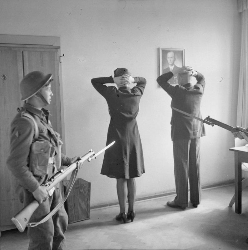 A British soldier with fixed bayonet guards two people with hands on their heads facing a wall on which a portrait hangs