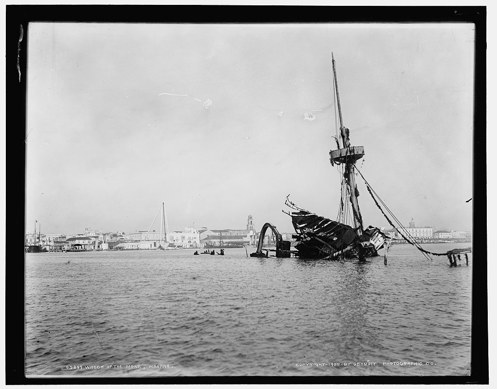 Photograph of the wrecked Maine in Havana harbour, a mast and twisted superstructure above the water, the city behind