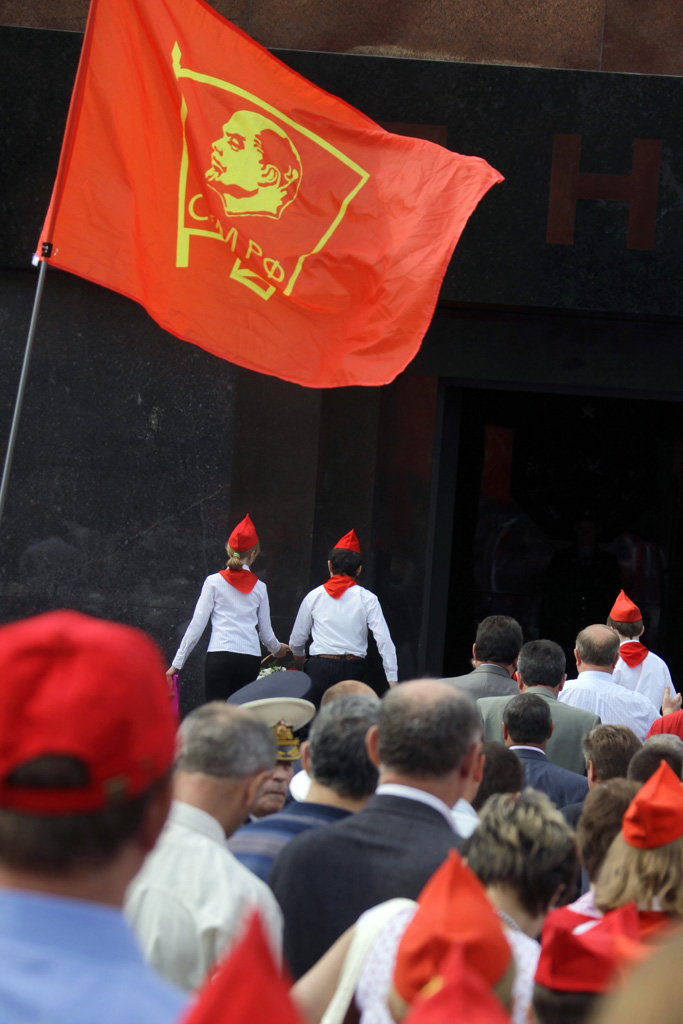 Photograph: a Young Pioneer neckerchief ceremony beside the black granite of Lenin's Mausoleum, under a red flag with Lenin's profile