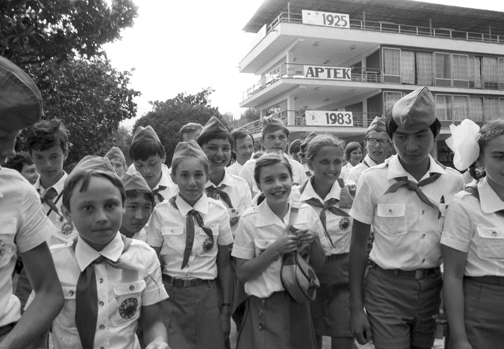 Photograph: a crowd of children in Pioneer uniform and neckerchiefs in front of a building with banners reading 1925, Artek and 1983