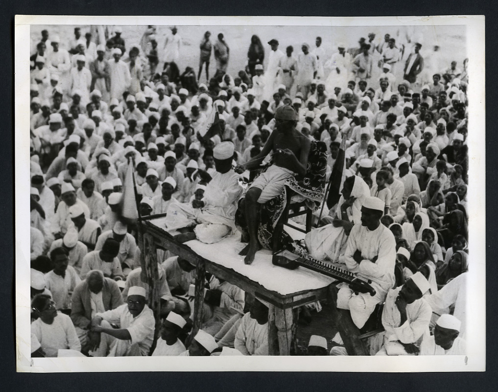 Gandhi spinning by hand on a raised platform while addressing a crowd, 1930