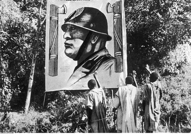 Three Ethiopian men raise the arm before a banner portrait of Mussolini in a steel helmet, Mekelle, November 1935