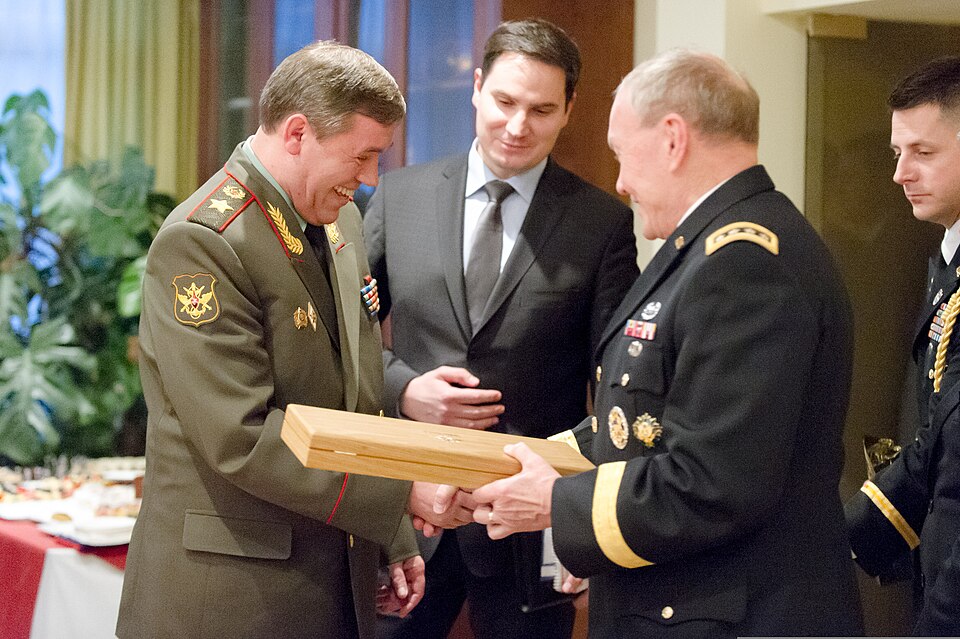 Russian General Valery Gerasimov in green service uniform, laughing and handing a wooden box to U.S. General Martin Dempsey in dark uniform