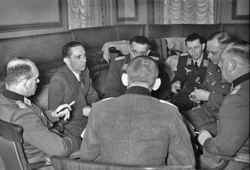 Joseph Goebbels in a civilian suit talking with Wehrmacht officers seated around a table in a Berlin hotel lounge