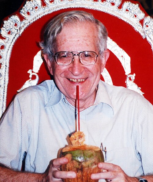 Noam Chomsky, smiling, holding a coconut drink with a straw, seated before a red and white ornate chair back