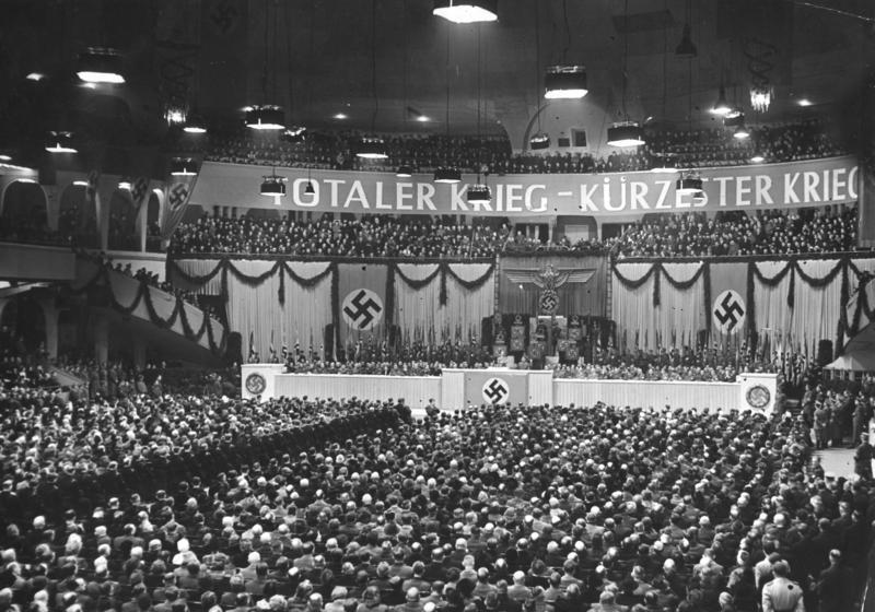 A packed Berlin Sportpalast hall with swastika banners and a stage banner reading "Totaler Krieg - Kürzester Krieg"