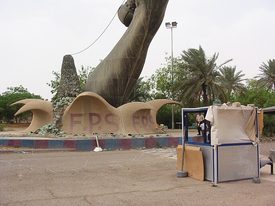 The base of one arm of the Victory Arch in Baghdad, with helmets set into the base, a net behind them and a small guard booth on the right