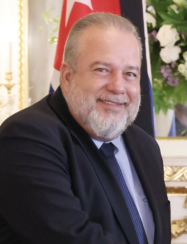 Manuel Marrero Cruz smiling, in a dark suit with a Cuban flag behind him, 28 September 2022