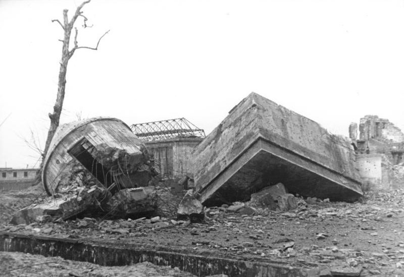 A tilted concrete slab and a toppled round concrete tower lying in a field of rubble, with a bare tree and a gutted building behind