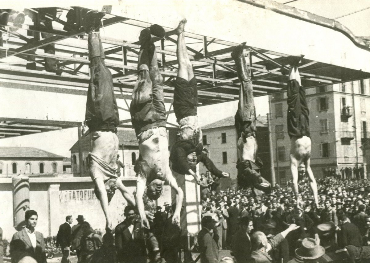 Several bodies hanging upside down by the feet from a steel girder frame above a crowd of onlookers in a Milan square