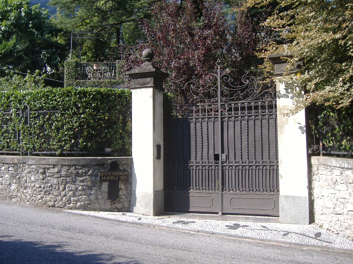 A dark iron gate between two white gateposts in a stone wall, with a small black cross plaque on the wall reading Benito Mussolini 28 aprile 1945