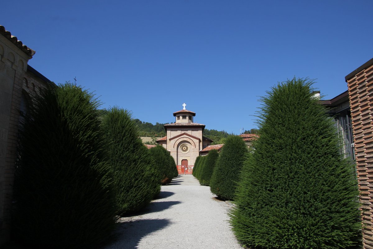A gravel path between tall clipped cypresses leading to a small brick chapel with a white cross on its roof, under a blue sky