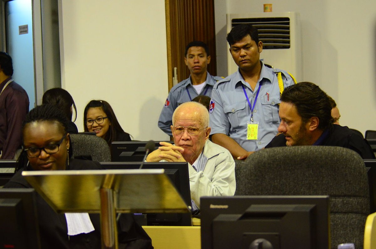 A white-haired man in glasses sits with his hands clasped in a courtroom, a uniformed guard standing behind him and lawyers in the foreground