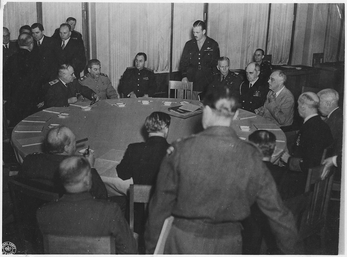 Delegates seated around a large round table in a curtained room, with Stalin at the far side among Soviet officers and Roosevelt in a light jacket at the right, a US Army officer standing behind them