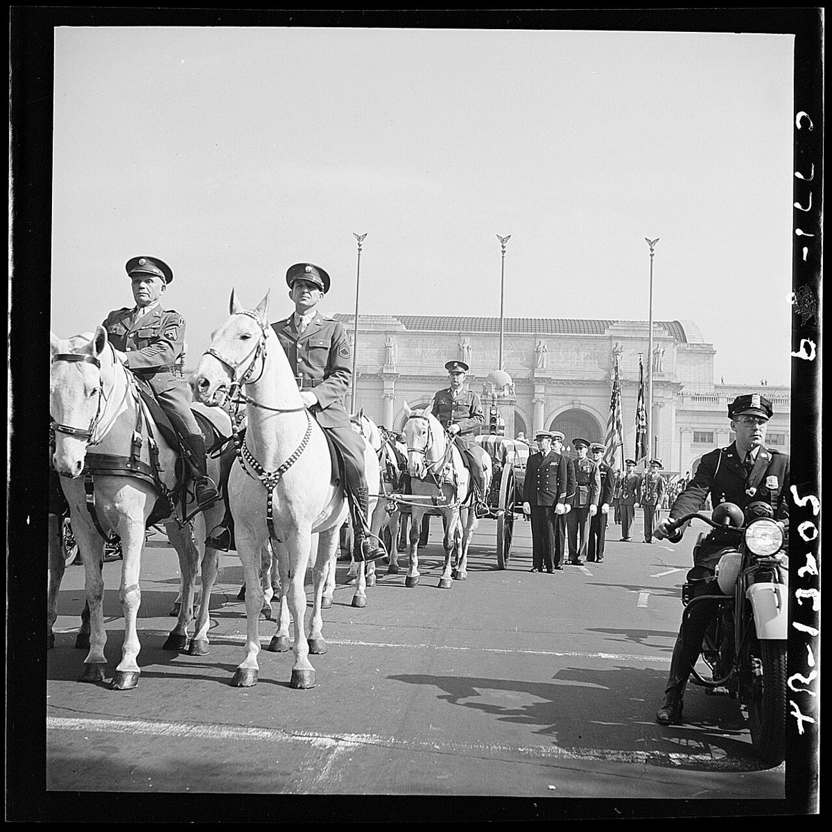 Two mounted soldiers on white horses in the foreground and a horse-drawn caisson with a flag-draped casket behind them, with honour guards of the services in front of Union Station, Washington