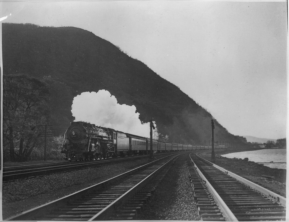 A steam locomotive pulling a long train along a riverside track beneath a wooded hill, white steam rising from the engine