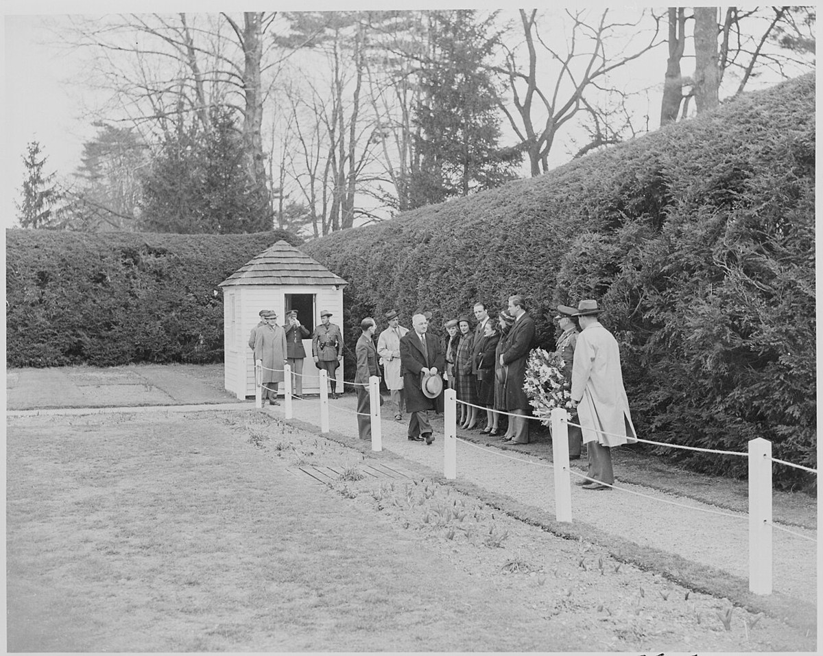 President Truman in a dark overcoat and holding his hat, walking along a roped gravel path beside a tall hedge, followed by a group with a wreath, a small white shed behind them