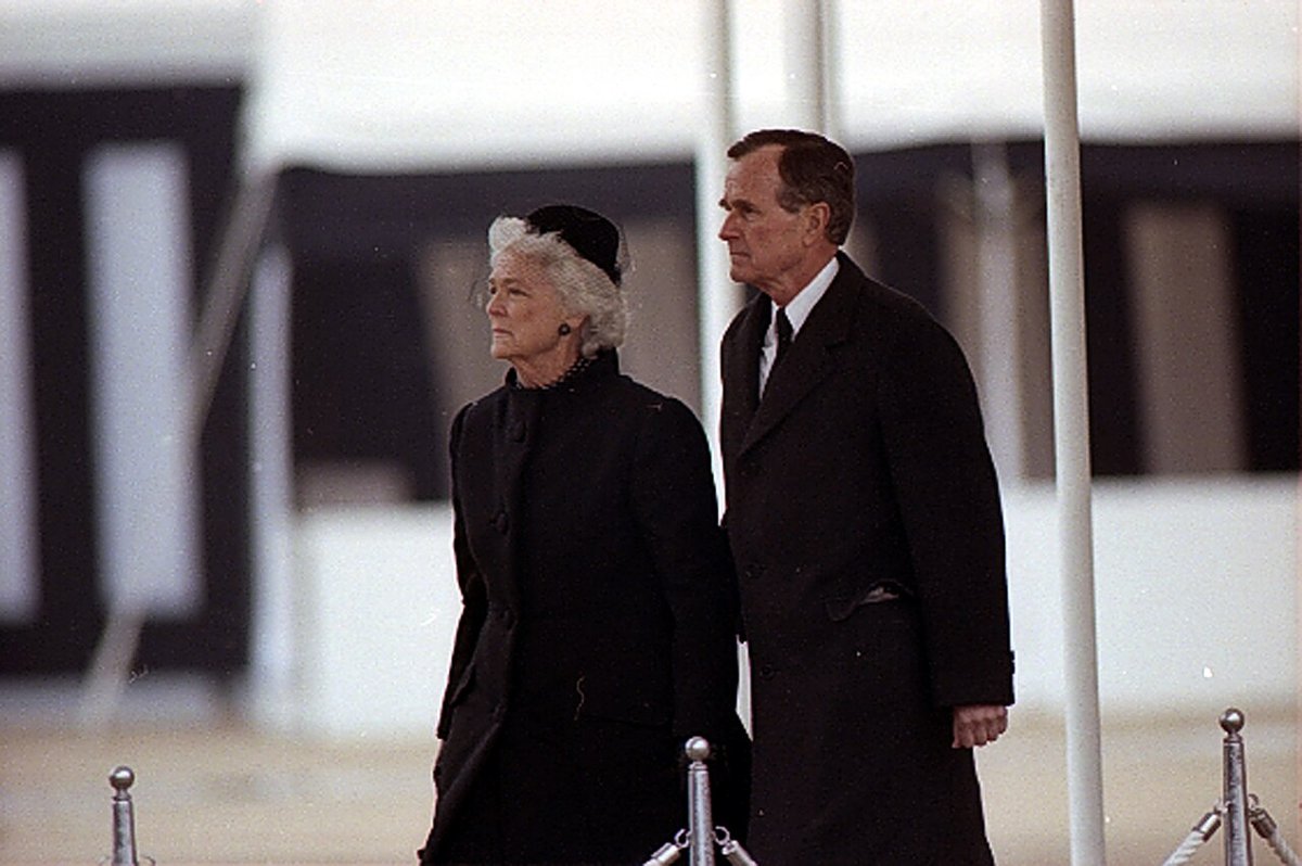 A grey-haired woman in a black coat and hat and a man in a dark overcoat stand side by side in the rain before a white and black funeral tent