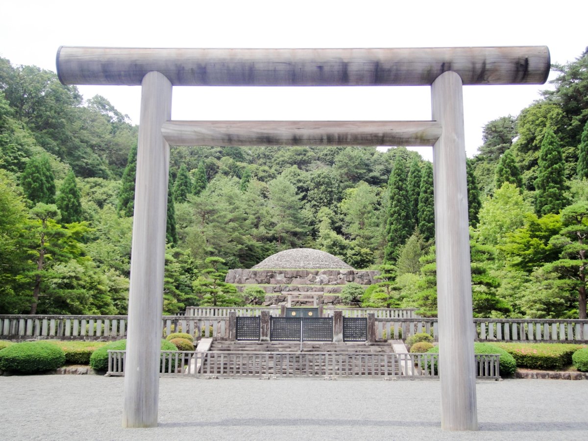 A wooden torii gate in the foreground frames a gated enclosure and a low stone-walled domed mound among trees