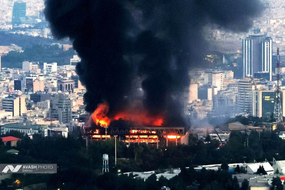 A multi-storey building burning at dusk under a large column of black smoke, with the Tehran skyline behind it