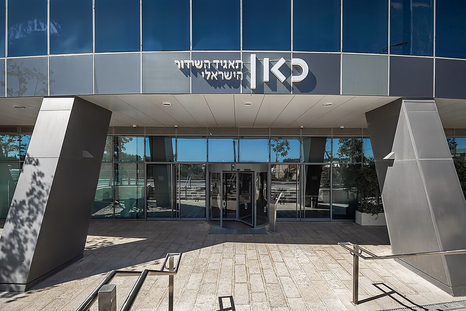 The glass-fronted entrance of a building under a sign reading, in Hebrew, 'Israeli Broadcasting Corporation' beside the Kan logo