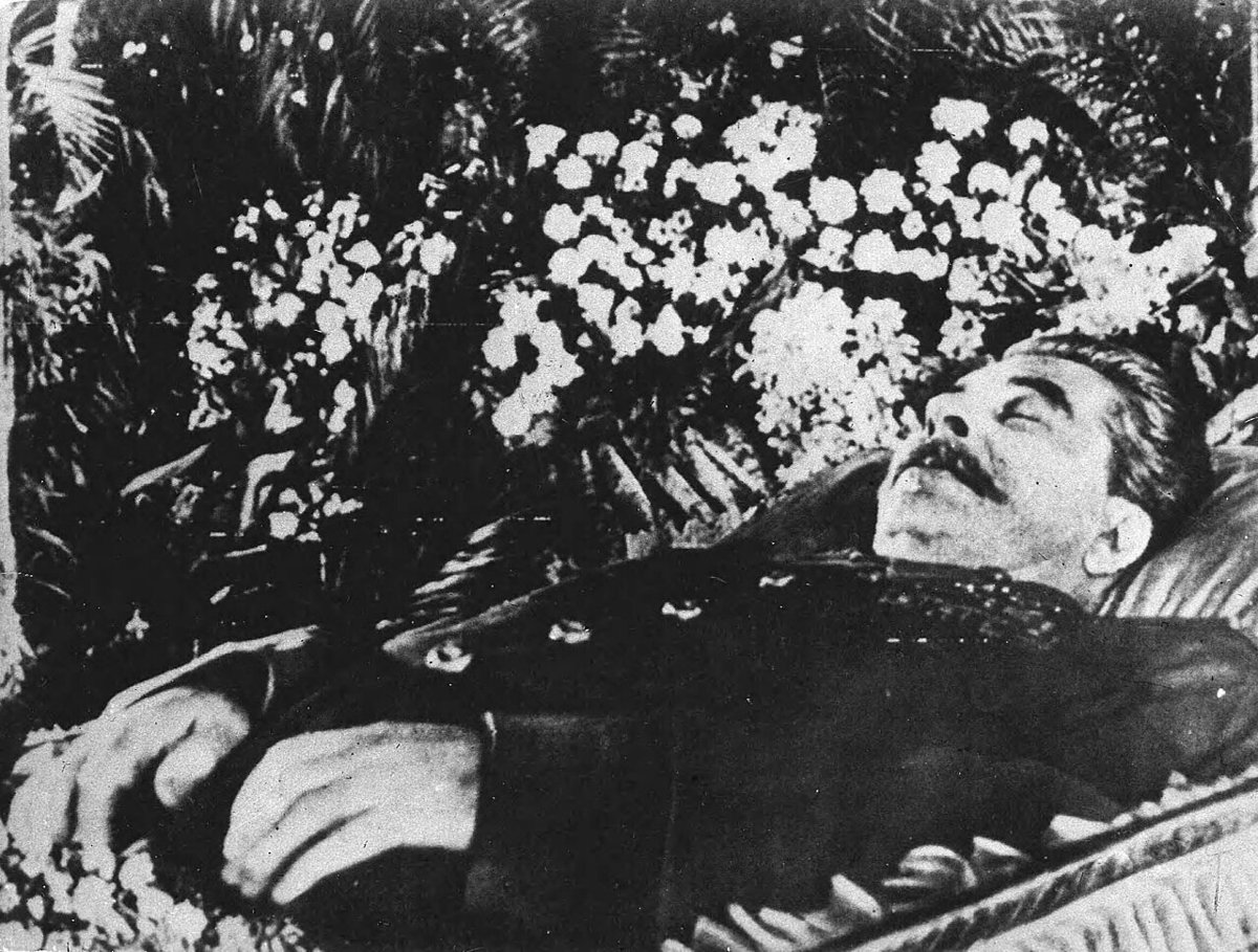 Black-and-white photograph of Stalin's head and shoulders lying in an open coffin, his eyes closed, among banks of flowers