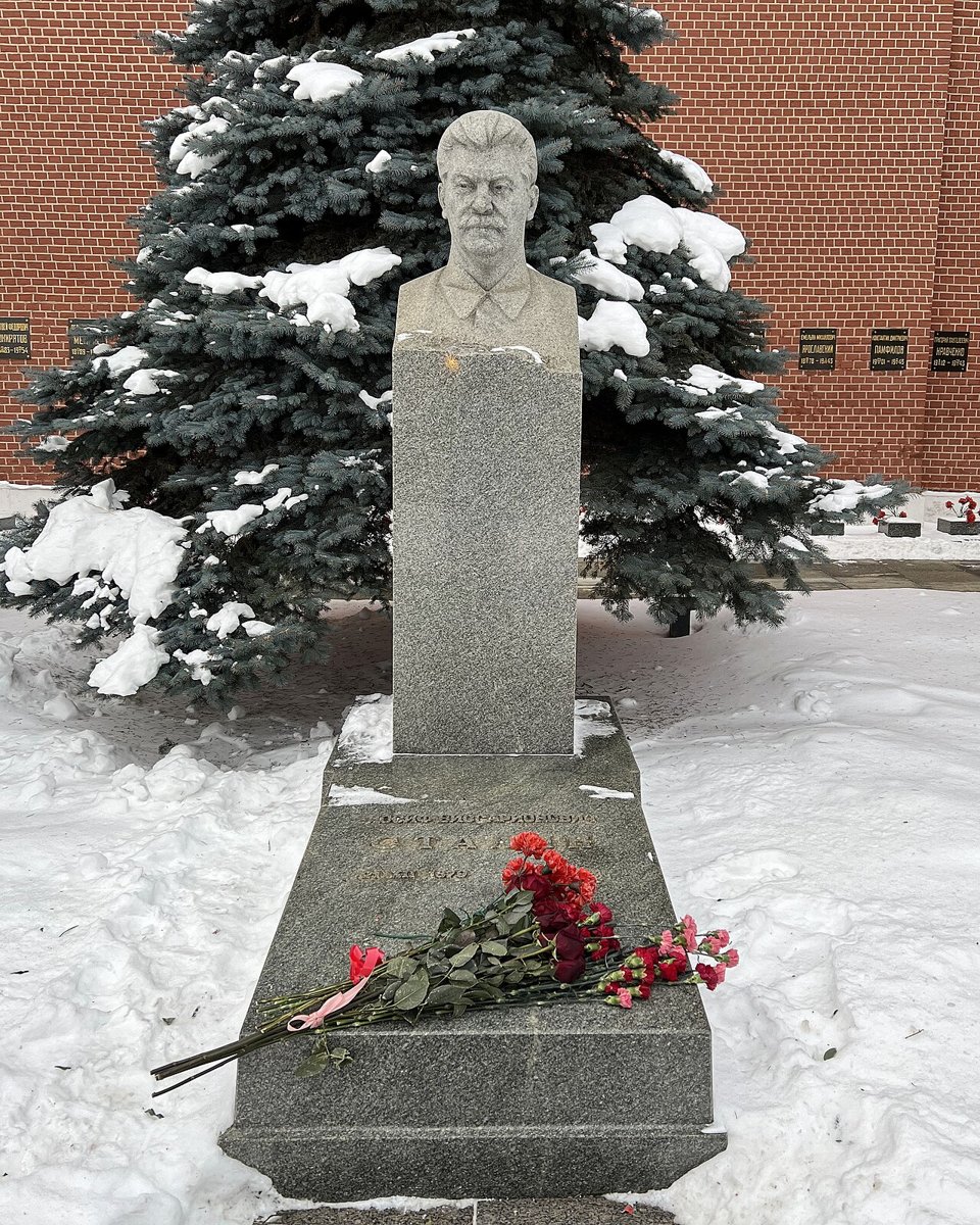 A granite grave marker with a bust of a man with a moustache, in front of a spruce tree and the red brick of the Kremlin Wall, with carnations laid on the slab in snow