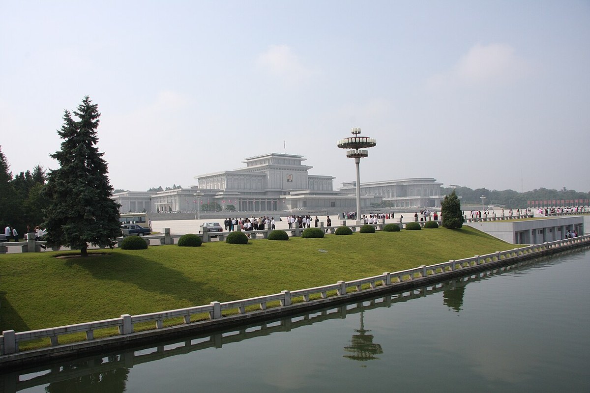 A wide white palace building behind a lawn and a canal, with a tall lamp tower to its right and visitors gathered in front