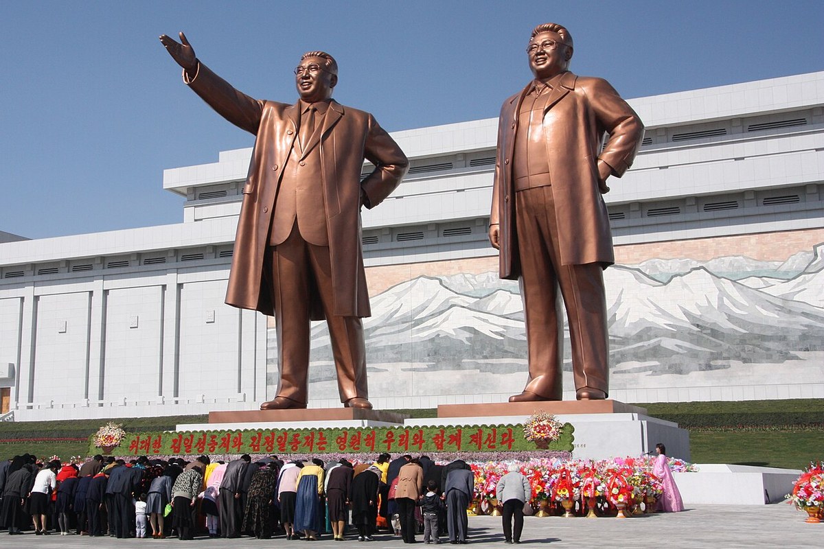 Two large bronze statues of standing men in long coats before a white building with a mosaic of a snowy mountain, with a crowd bowing in front and flower baskets at the base