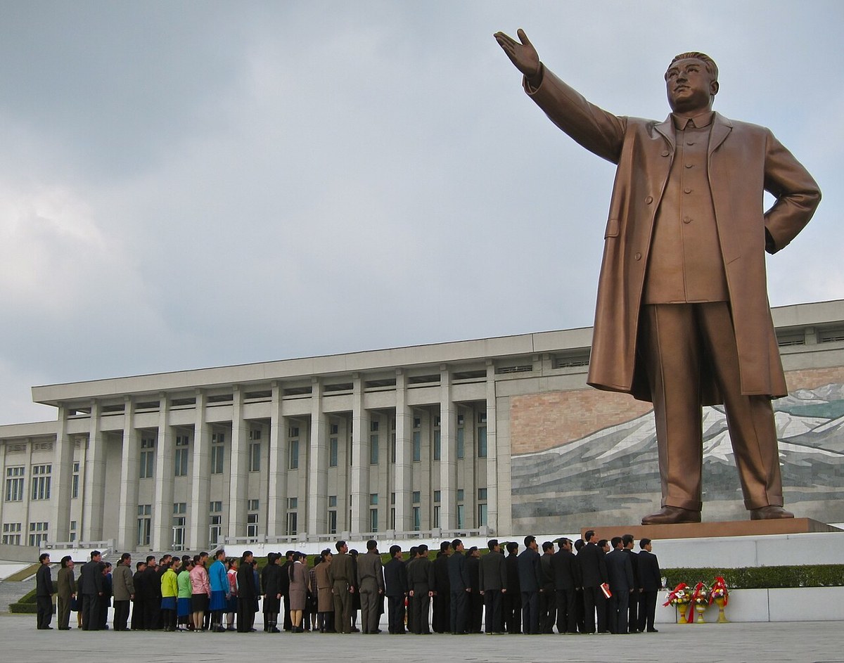 A bronze statue of Kim Il Sung with one arm raised above a line of visitors standing before a colonnaded building