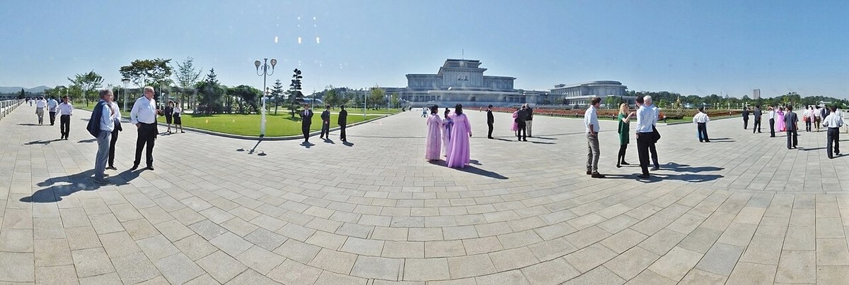 A wide panoramic view across a large paved plaza with scattered visitors toward a distant white palace building