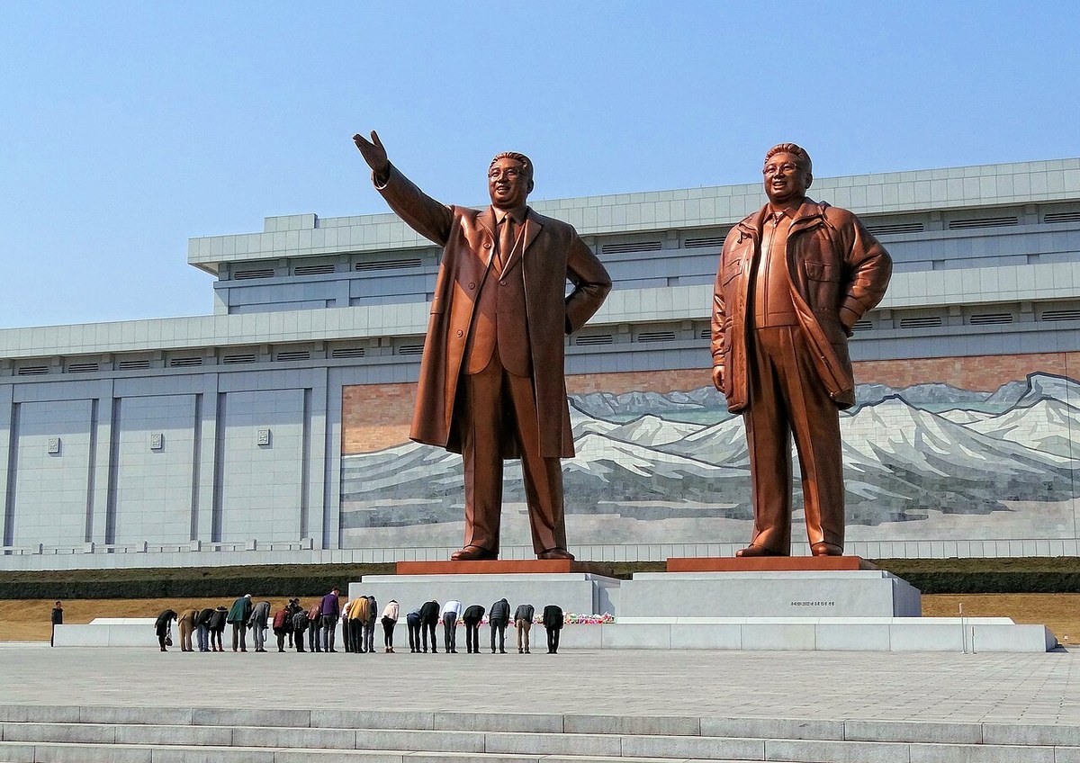 Two large bronze statues of men in long coats before a white building with a mosaic of snowy mountains, with a row of visitors bowing in front