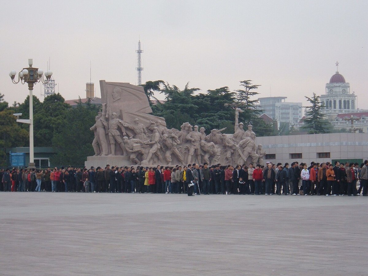 A long line of people standing in front of a stone sculpture group of figures with a flag, in Tiananmen Square