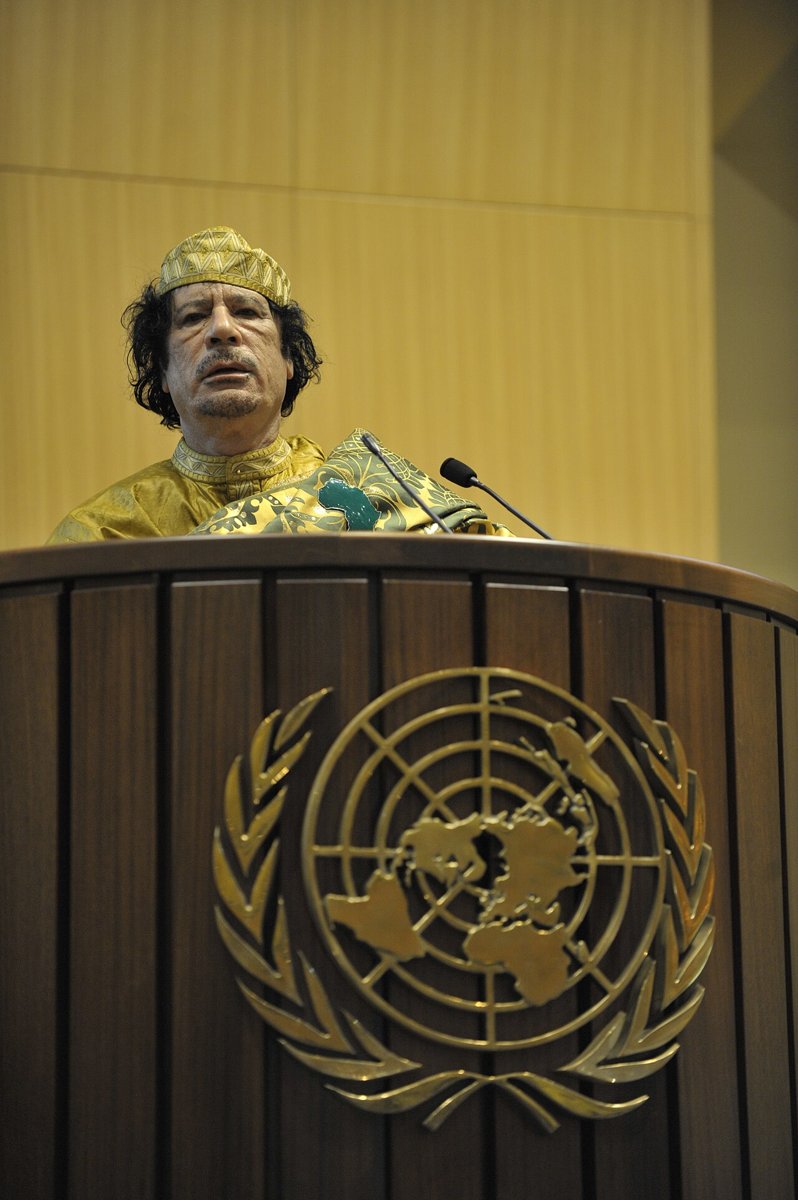 Muammar Gaddafi in a gold embroidered robe and cap speaking at a wooden lectern bearing the United Nations emblem