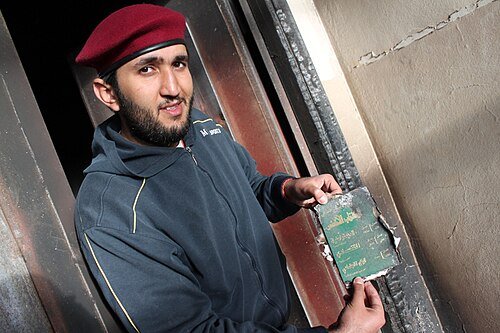 A man in a red beret and a dark hooded top holds up the burnt cover of a small green book beside a charred doorway